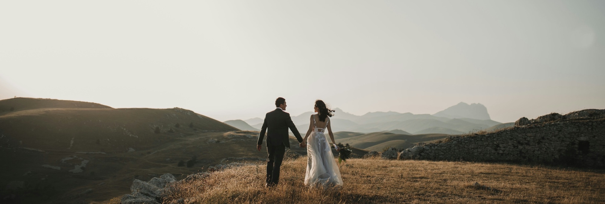 A guest's wedding photo at full width — a couple walking a golden hillside at sunset, mountain ridges behind them and a dry stone wall to the right