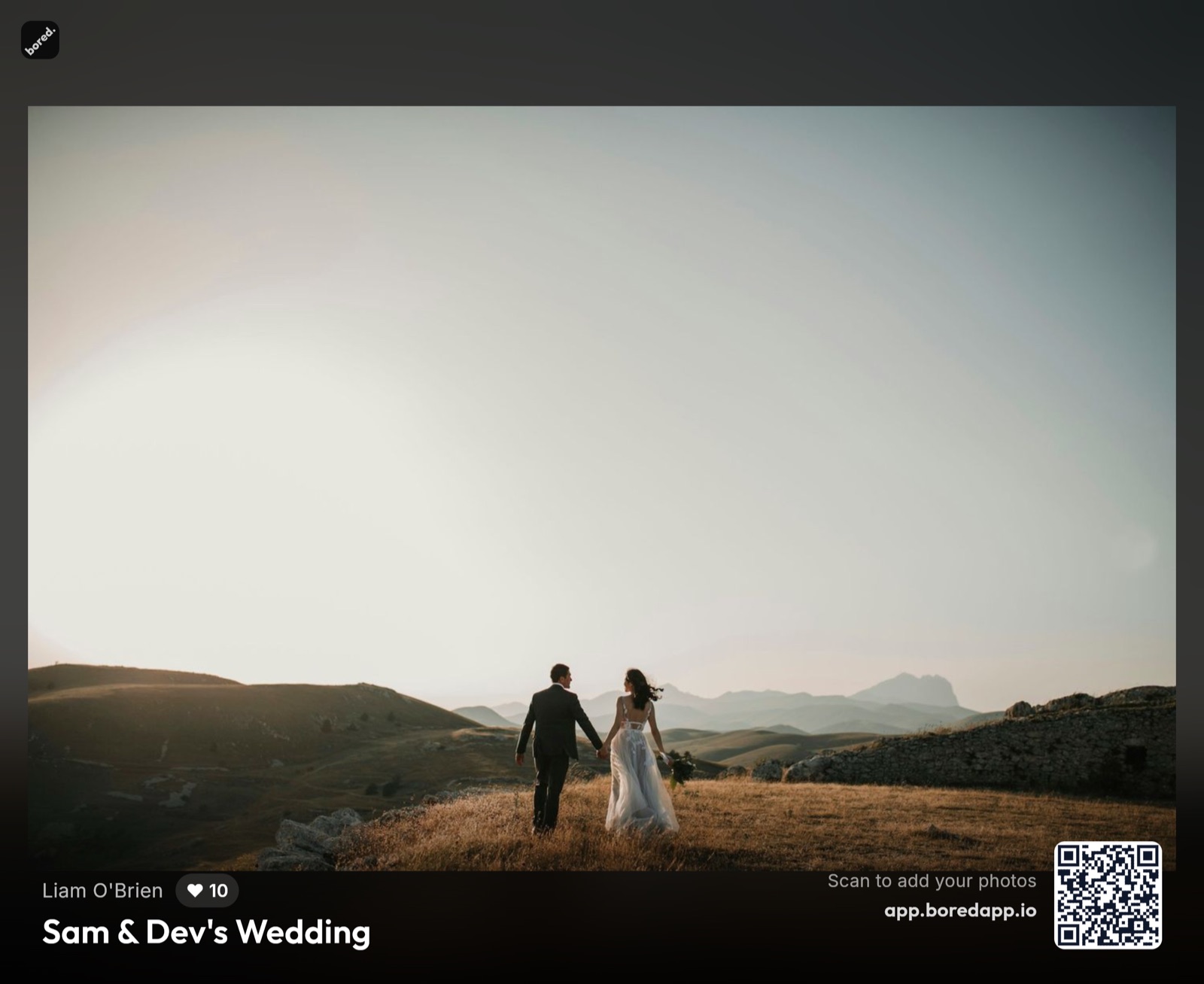 A BoredApp Photo Wall on a venue screen at a wedding — a guest's photo of the couple walking a golden hillside at sunset filling the frame, the guest's name, a heart count, and the event title along the bottom, and a QR code captioned Scan to add your photos