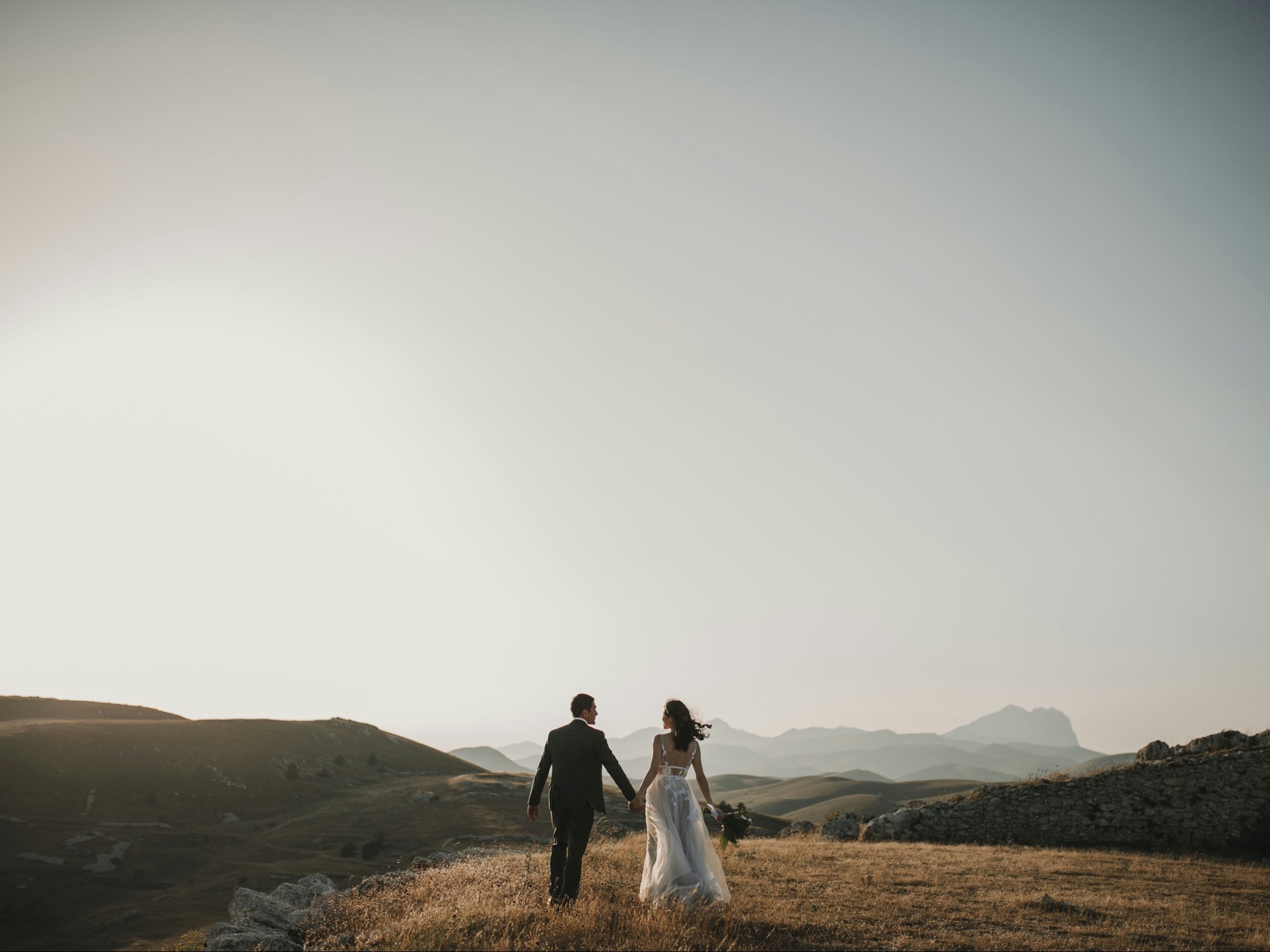 A guest's wedding photo at full size — a couple walking a golden hillside at sunset, mountain ridges behind them under a wide pale sky